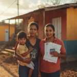 Família brasileira em frente a casa simples segurando cartão do Bolsa Família e documentos do Cadastro Único em cidade do interior